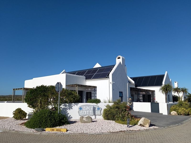 A well-maintained white coastal-style house featuring solar panels on the roof, a paved driveway, and landscaped garden beds.