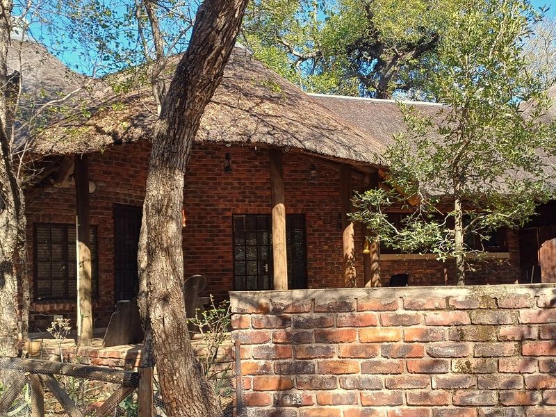 A rustic exterior view of a house featuring a traditional thatch roof, exposed brick walls, and timber support columns set within a natural garden.