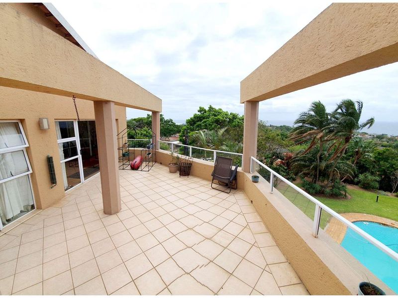 Spacious elevated balcony featuring tiled flooring, glass balustrades, and structural pillars, offering scenic views over the garden and swimming pool area.