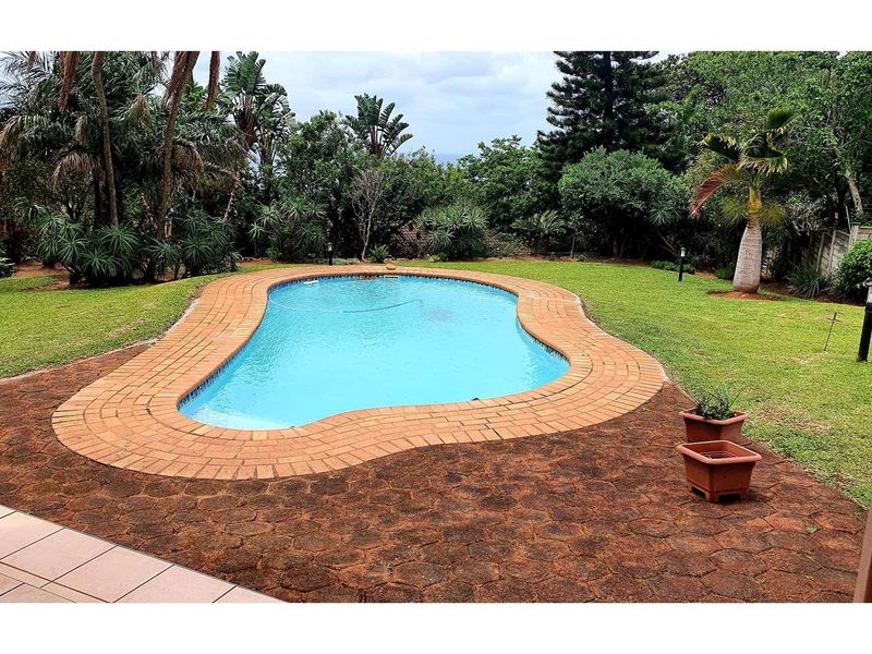 A well-maintained swimming pool set within a lush, landscaped garden featuring brick coping and a paved patio area.