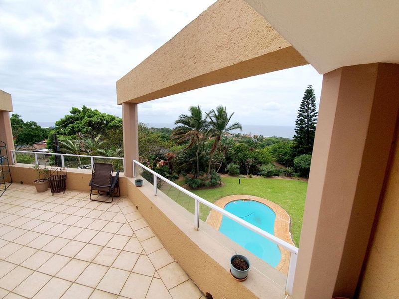 Spacious elevated balcony featuring tiled flooring and a glass balustrade, offering scenic views over the garden and swimming pool area.