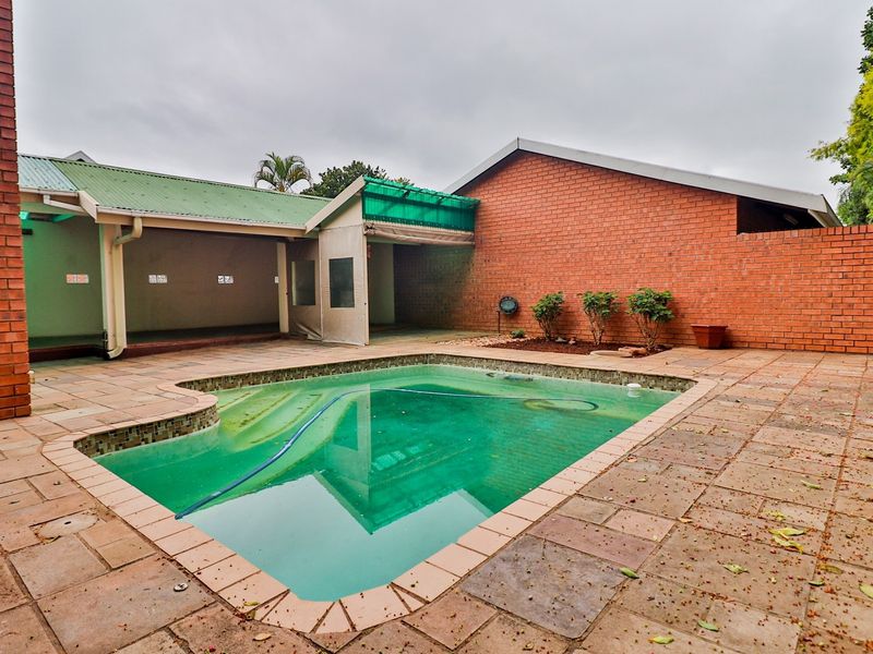 Outdoor pool area featuring a tiled surround, brick boundary walls, and a covered patio space, requiring maintenance to the pool water and surrounding surfaces.