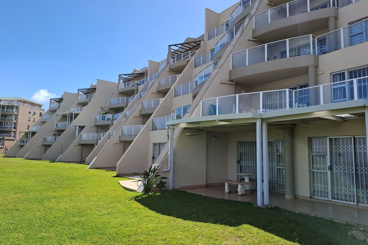 Exterior view of a multi-story apartment complex featuring stepped balconies with glass railings and ground-level patios overlooking a landscaped lawn.