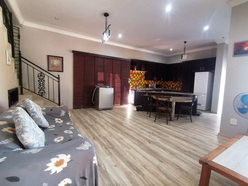 Open-plan living area featuring laminate flooring, a staircase with wrought iron railing, and a kitchen with dark cabinetry and a tiled backsplash.