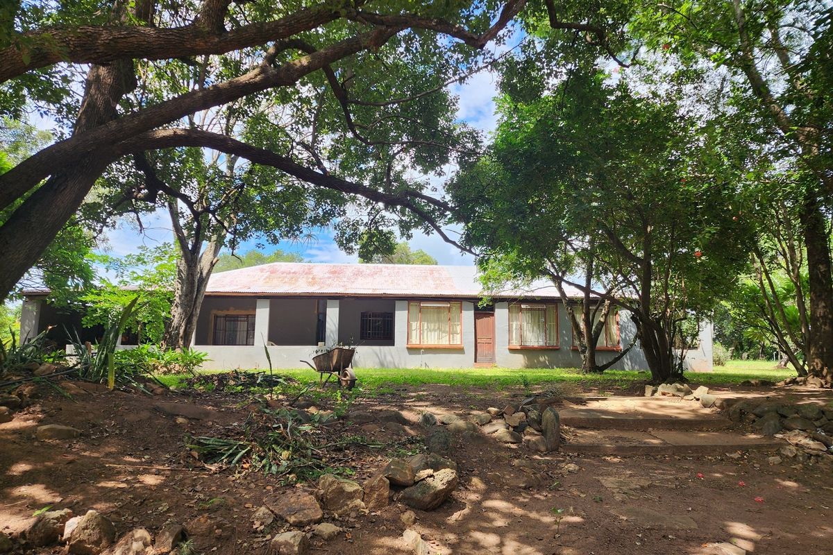 Exterior view of a farm-style dwelling featuring a corrugated metal roof, set within a mature, tree-lined garden landscape.