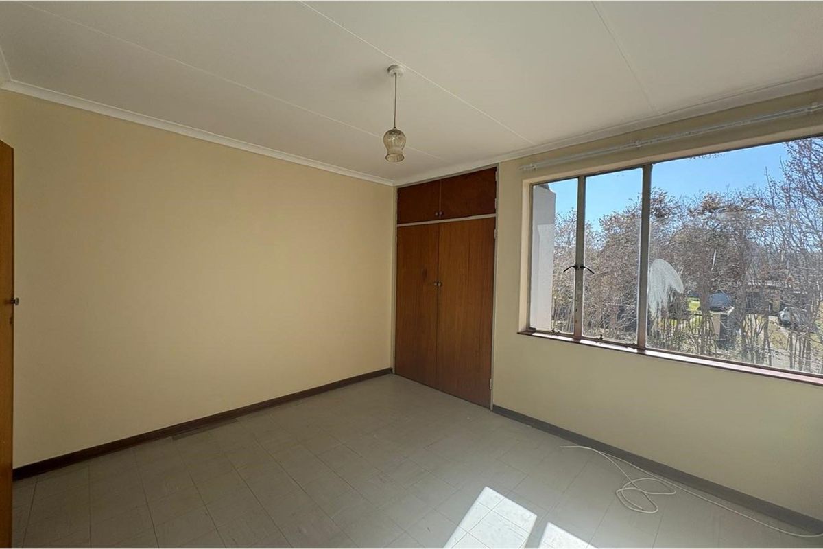 A vacant bedroom featuring built-in wooden cupboards, tiled flooring, and large windows providing natural light and an exterior view.