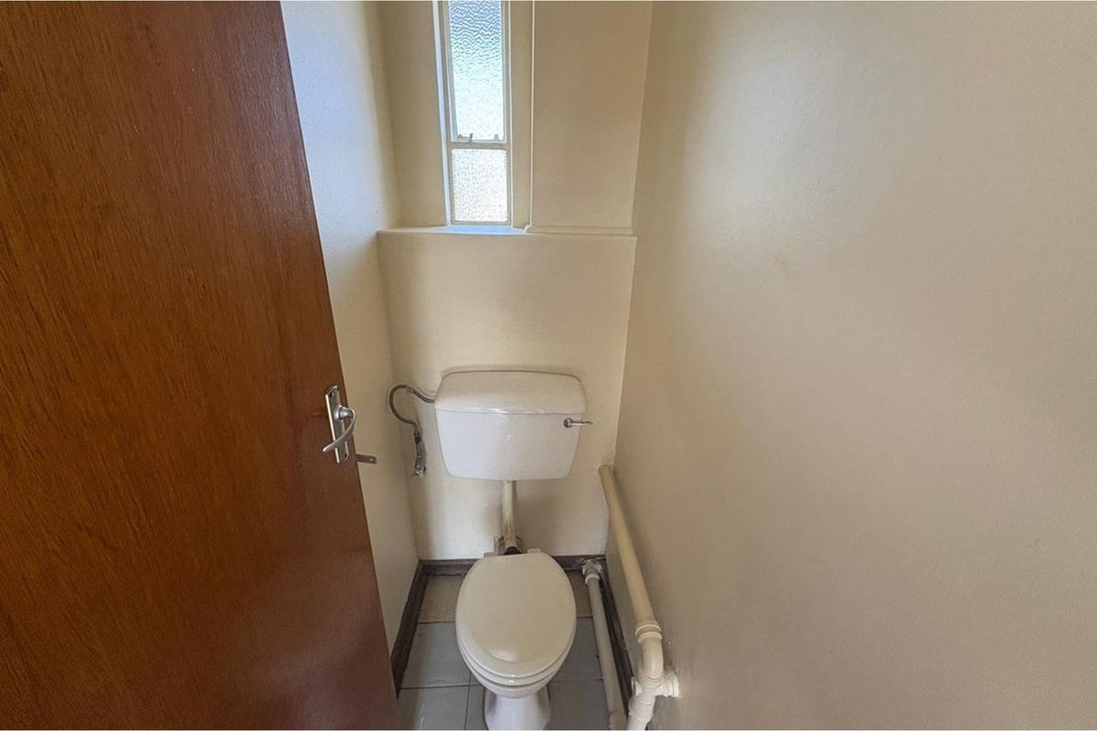 A compact, dated bathroom featuring a standard toilet, exposed plumbing, and a small frosted window.