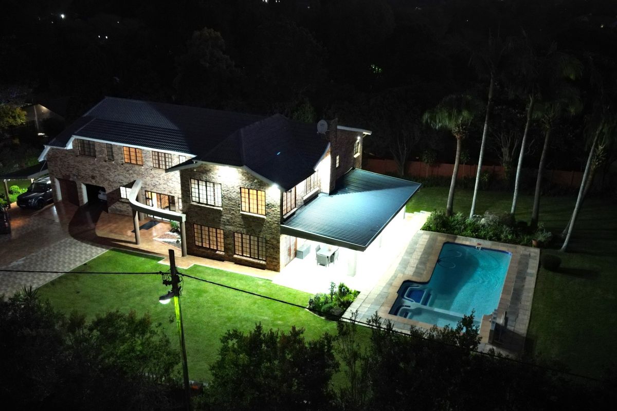 Aerial night view of a double-story brick house featuring a swimming pool, covered patio, and landscaped garden.