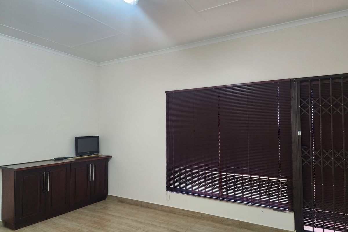 A living area featuring tiled flooring, built-in wooden cabinetry, and a window fitted with dark horizontal blinds and a security gate.