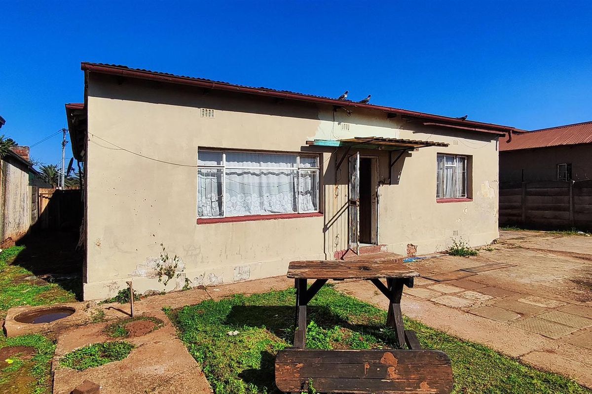 Rear view of a single-story house featuring a paved patio area, metal window frames, and a corrugated roof.
