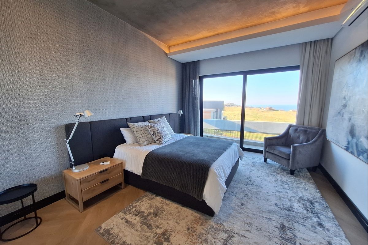 Modern bedroom featuring a textured feature wall, recessed ceiling lighting, herringbone flooring, and large sliding glass doors opening to a scenic coastal view.