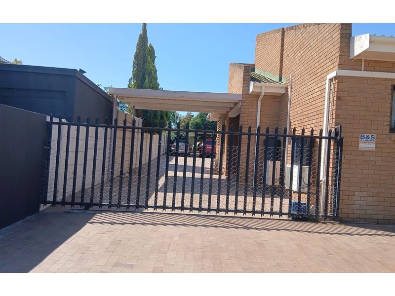 A secure driveway entrance featuring a black metal security gate, paved driveway, and a covered carport area alongside the brick residential building.