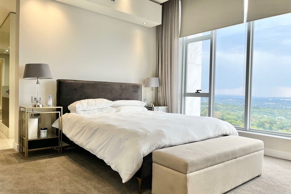 A modern, well-appointed bedroom featuring floor-to-ceiling windows with expansive views, neutral carpeting, and recessed ceiling lighting.