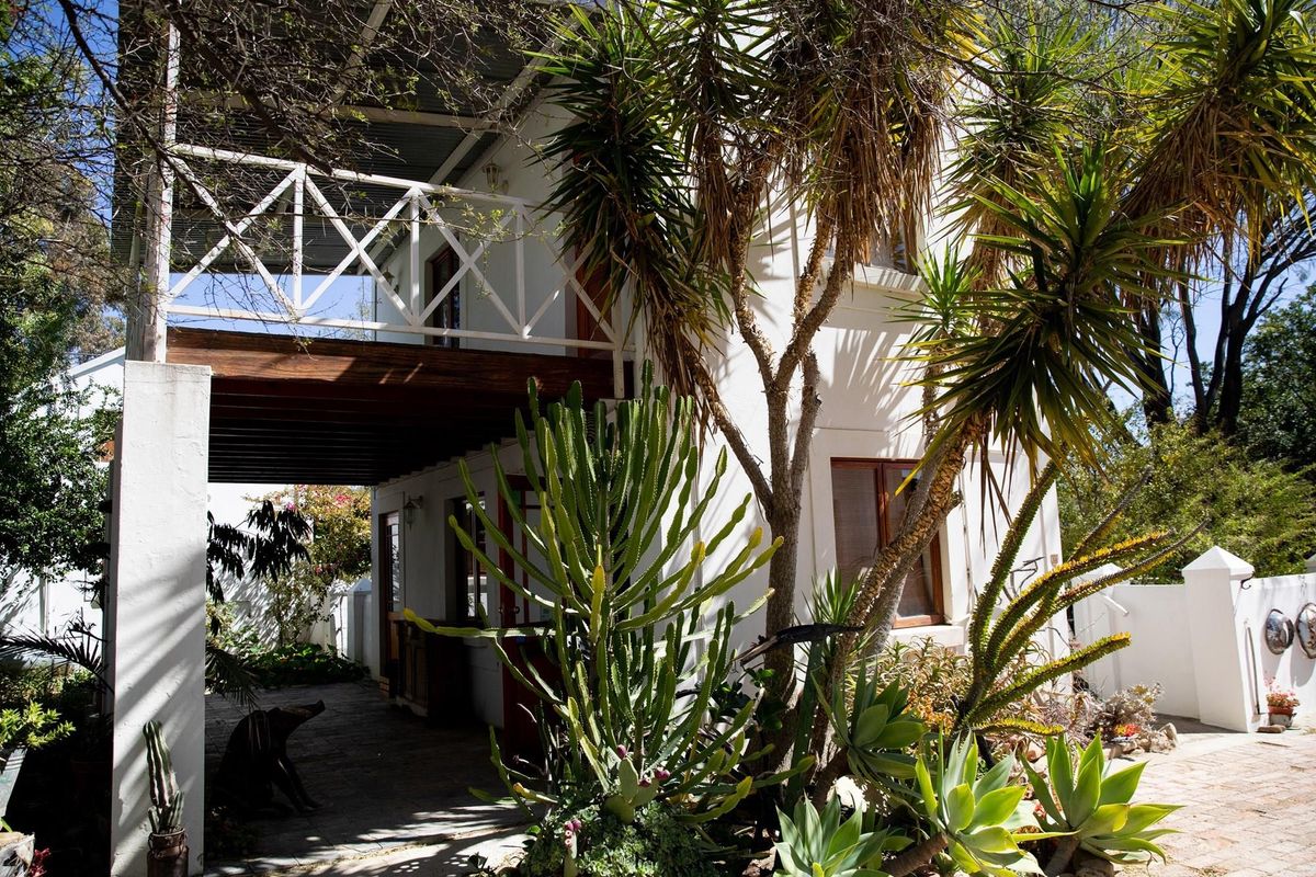 A two-story exterior view featuring a covered patio area and an upper-level balcony with timber railings, surrounded by established garden landscaping.