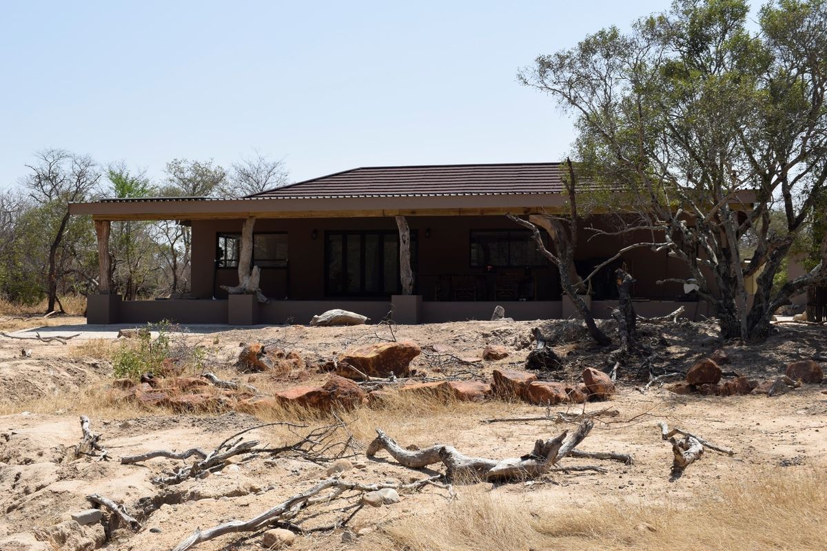 A rear view of a single-story house featuring a covered patio area with natural timber support posts, set within a dry, bushveld landscape.