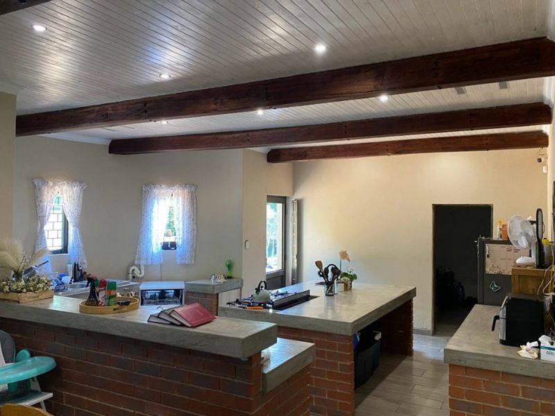 Spacious open-plan kitchen featuring exposed timber ceiling beams, concrete countertops, and brick base cabinetry, offering a rustic farmhouse aesthetic.