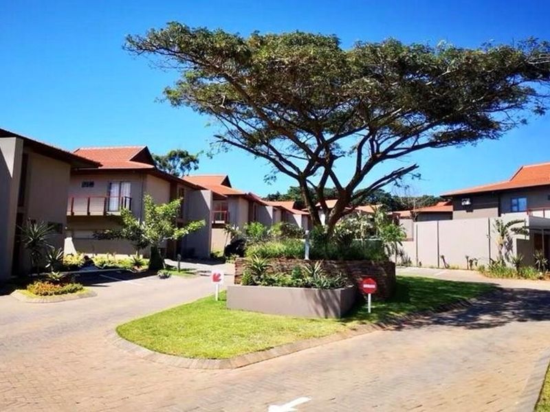 A view of the townhouse complex entrance featuring paved driveways, landscaped garden beds, and mature trees under a clear blue sky.