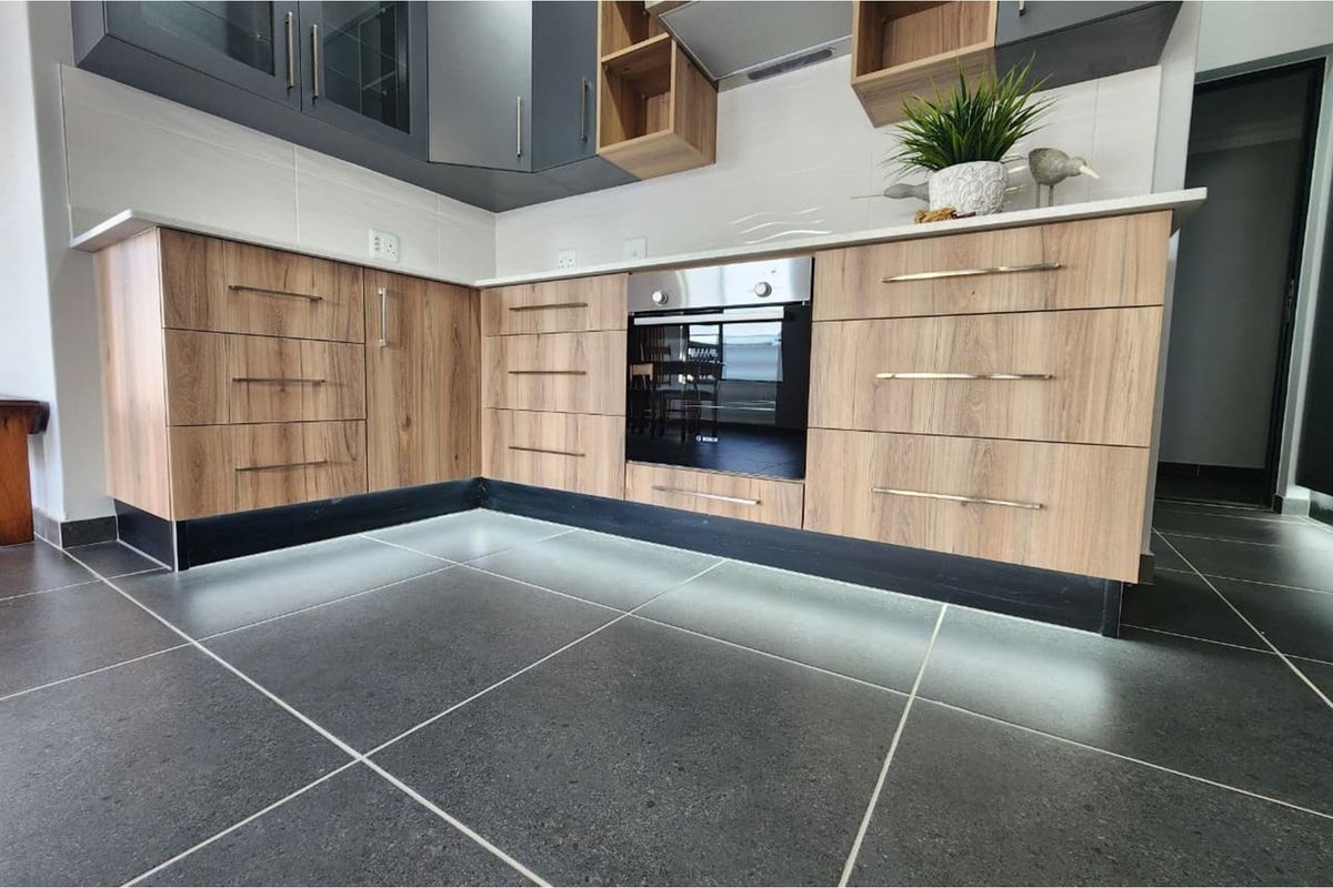 Modern kitchen featuring wood-grain cabinetry, integrated oven, sleek white countertops, and dark floor tiles with under-cabinet LED lighting.