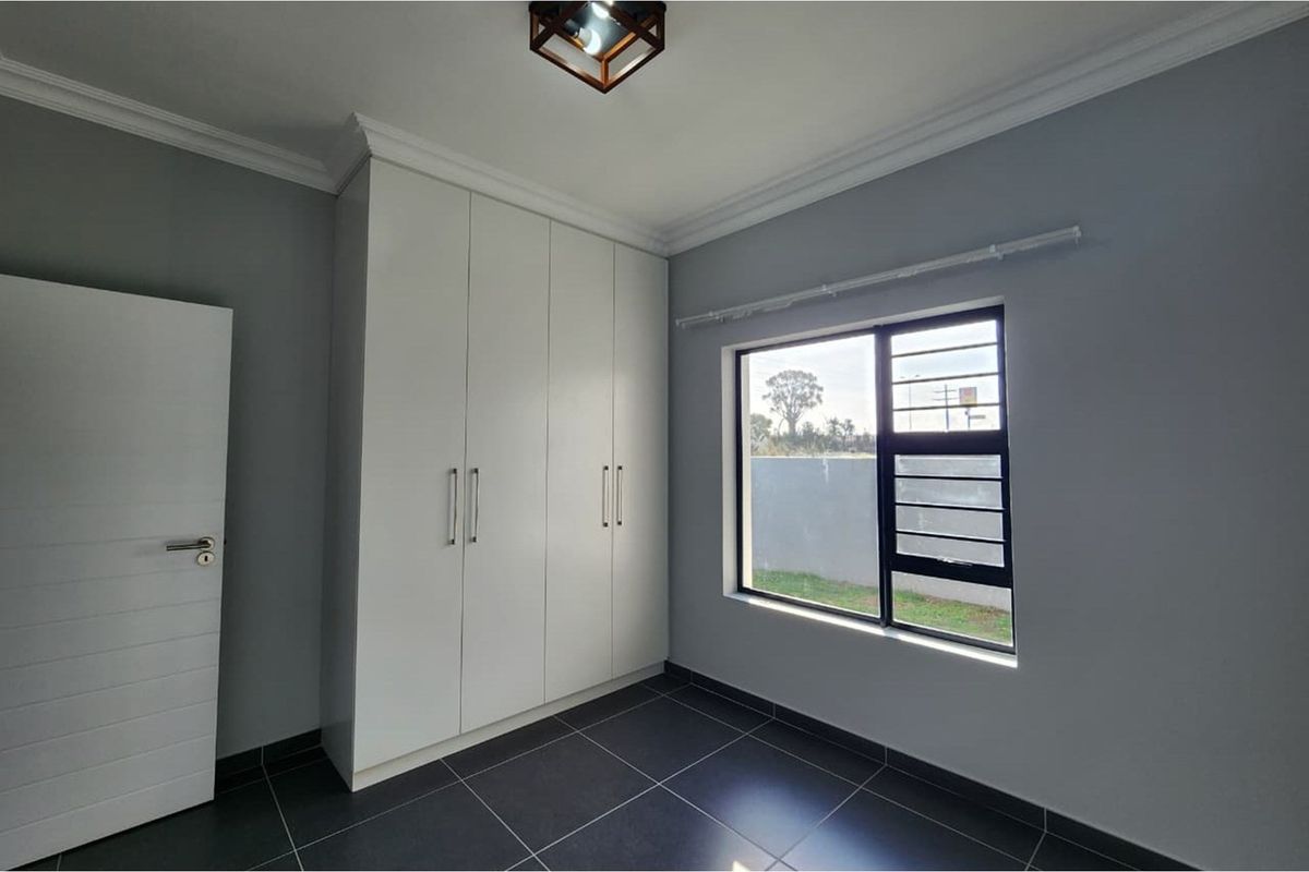 A clean, modern bedroom featuring built-in white cabinetry, dark floor tiles, and a window with security bars.
