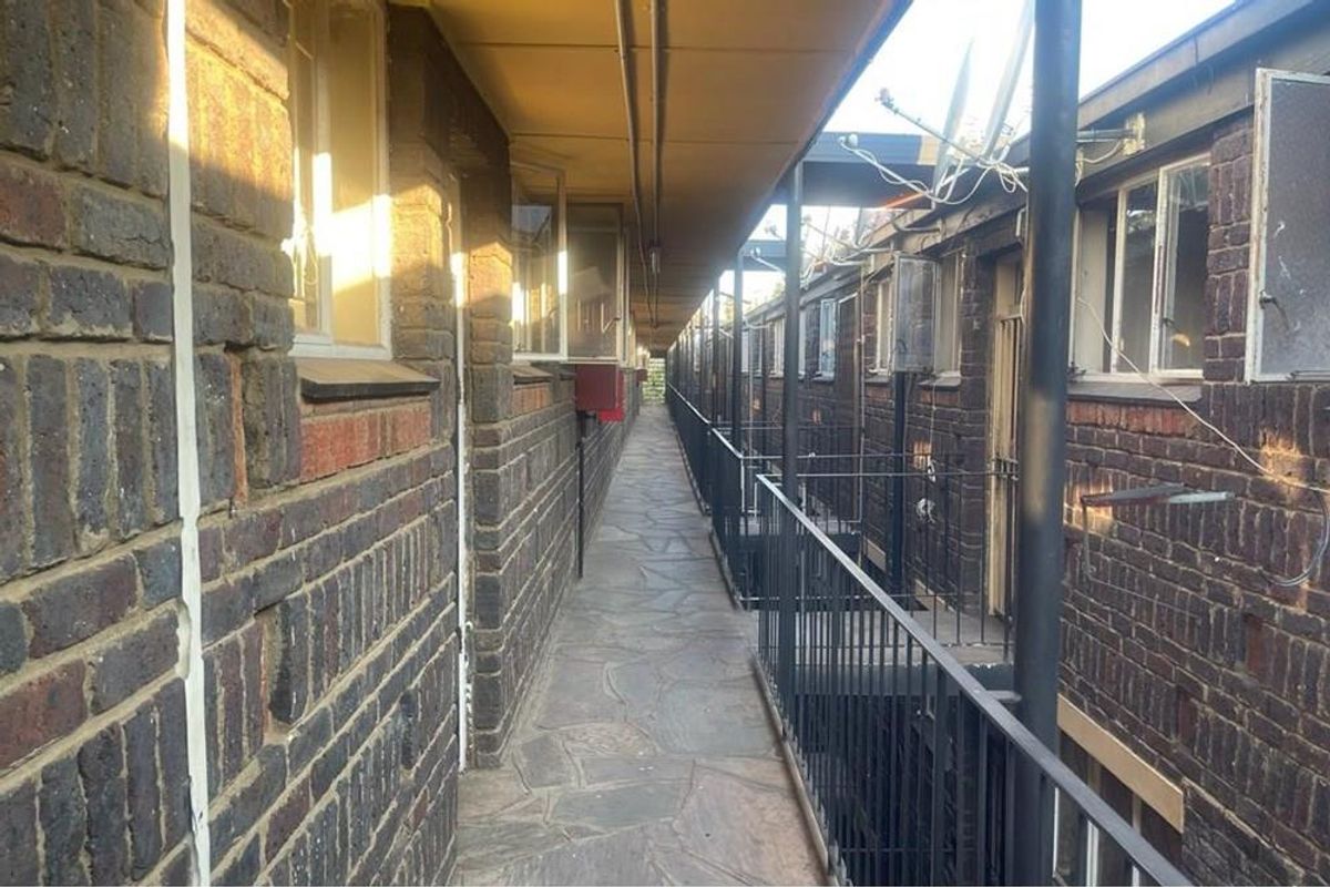 An exterior view of an apartment complex walkway featuring exposed brick walls, metal safety railings, and stone paved flooring.