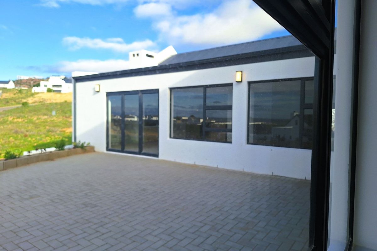 A view of the rear exterior patio featuring paved flooring, modern white facade, and large dark-framed windows and doors.