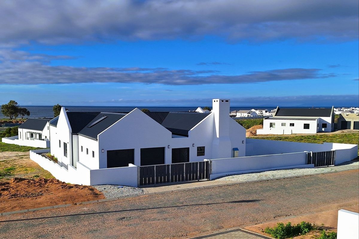 Aerial view of a modern white house with a dark metal roof, featuring a perimeter wall, gated entrance, and coastal views.