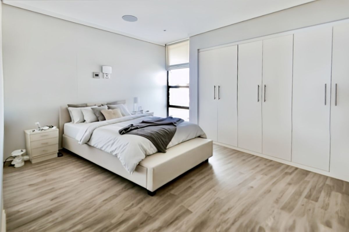 A modern, well-maintained bedroom featuring light laminate flooring, extensive built-in cupboards, and neutral finishes, creating a bright and comfortable living space.