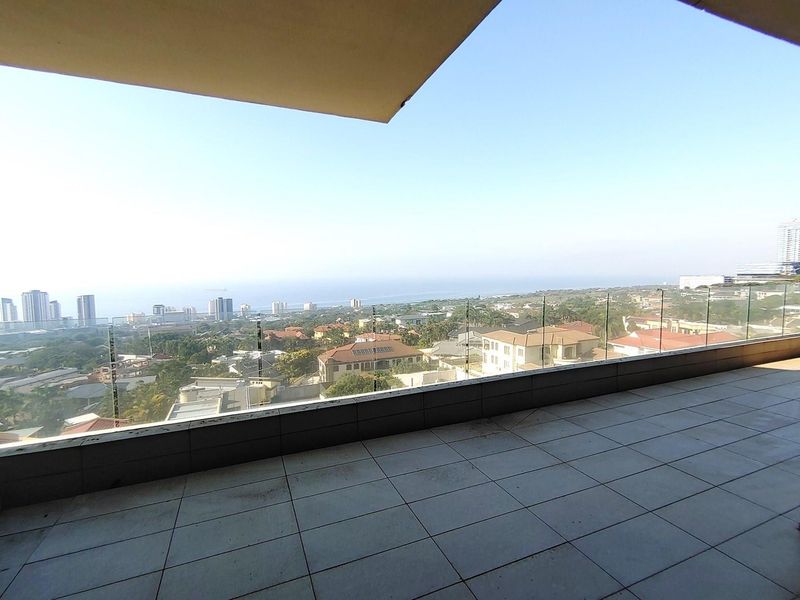 Spacious penthouse balcony featuring tiled flooring and a modern glass balustrade, offering expansive panoramic views of the surrounding cityscape and coastline.