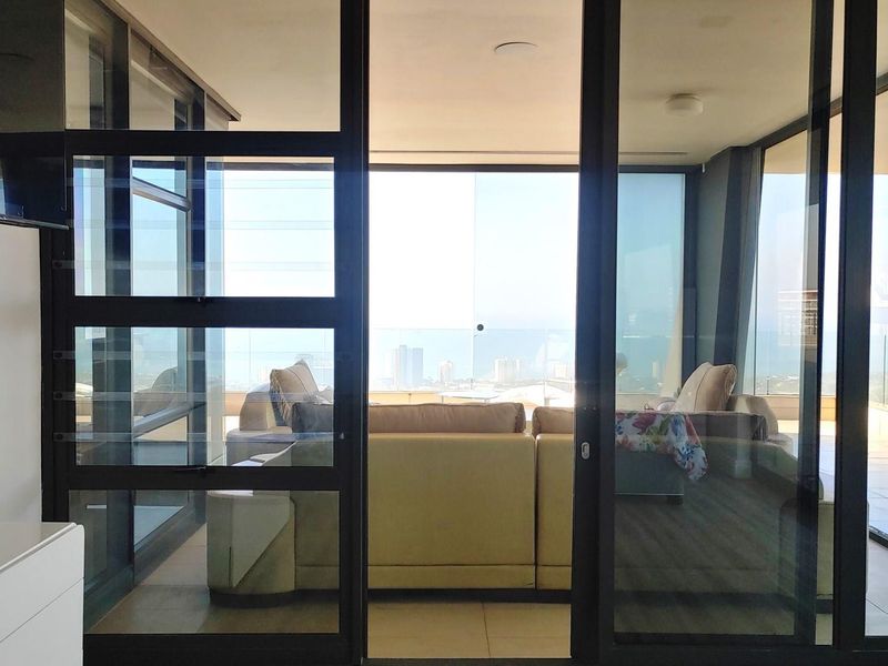 A spacious covered balcony featuring large glass sliding doors, tiled flooring, and a glass balustrade offering expansive city and coastal views.