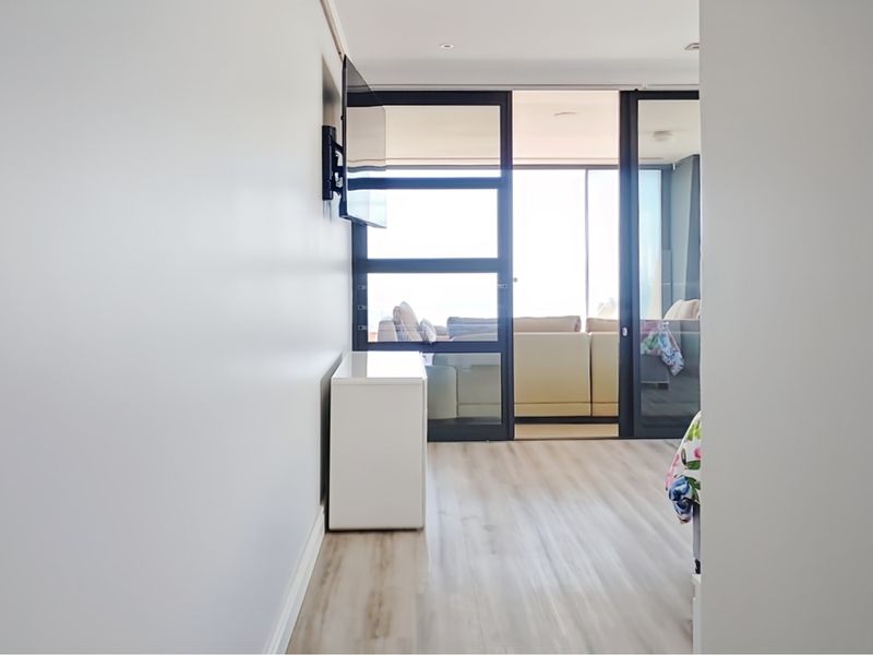 A bright hallway featuring light laminate flooring and recessed lighting, leading through black-framed glass doors into an adjacent living space.
