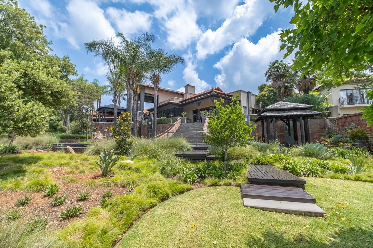 A beautifully landscaped rear garden featuring tiered lawns, mature trees, a wooden walkway, and a gazebo, leading up to the main residence via stone stairs.