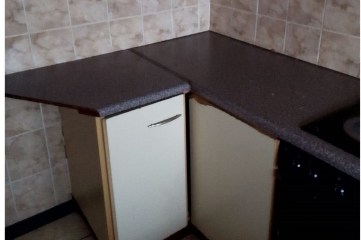 A compact kitchen corner featuring laminate countertops, built-in cabinetry, and ceramic wall tiling, showing signs of wear and tear on the cabinet finishes.