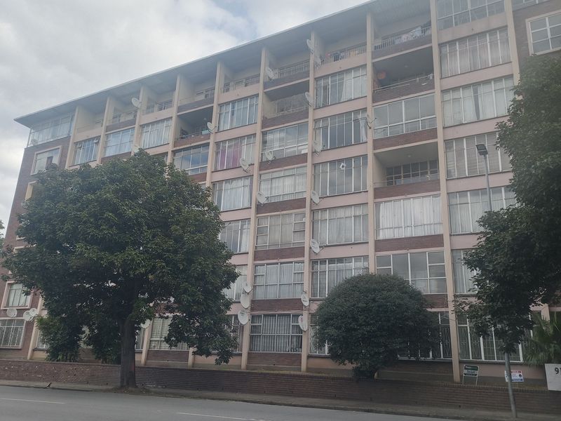 A multi-story apartment building featuring a brick facade, numerous balconies, and large windows, situated along a street.
