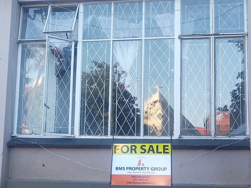 Exterior view of an apartment building facade featuring large casement windows with security burglar bars and a prominent for sale sign.