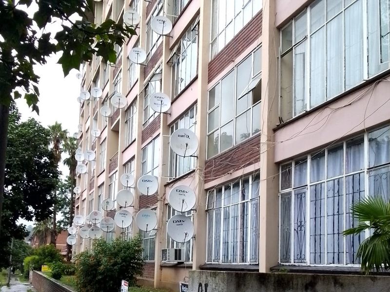 Exterior view of an apartment building facade featuring numerous satellite dishes and large windows, showing signs of wear and external wiring clutter.