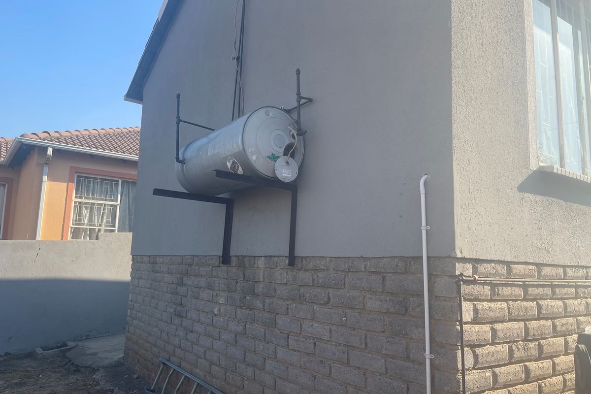 Exterior view of the property showing an externally mounted electric water heater cylinder with exposed plumbing and mounting brackets.
