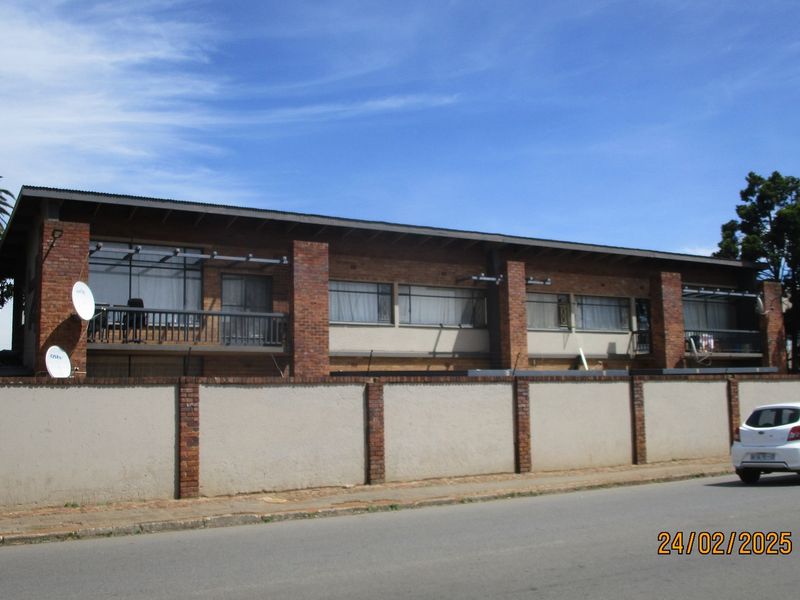 Street-facing view of a multi-story commercial building featuring brick facade, balconies with metal railings, and a surrounding perimeter wall.