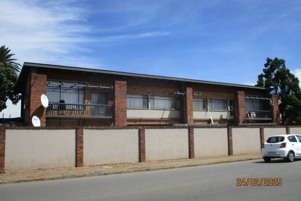 Street-facing view of a multi-story commercial building featuring brick facade, balconies with metal railings, and a surrounding perimeter wall.