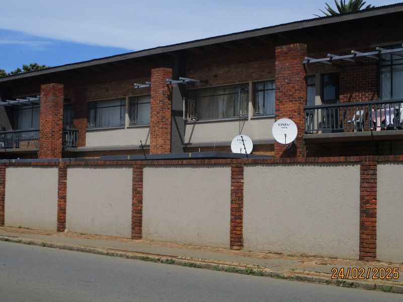 Street-facing view of a multi-unit commercial or residential building featuring brick facade, balconies with metal railings, and a perimeter boundary wall.