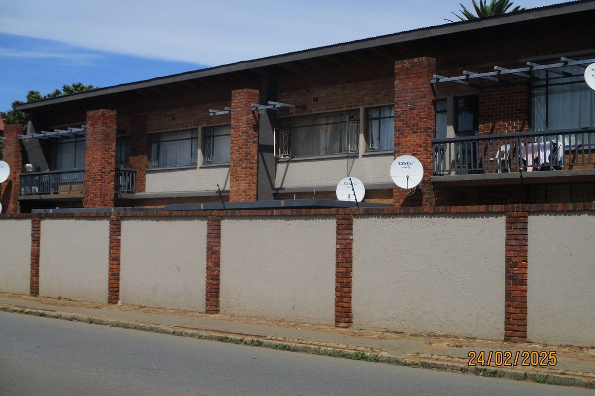 Street-facing view of a multi-unit commercial or residential building featuring brick facade, balconies with metal railings, and a perimeter boundary wall.