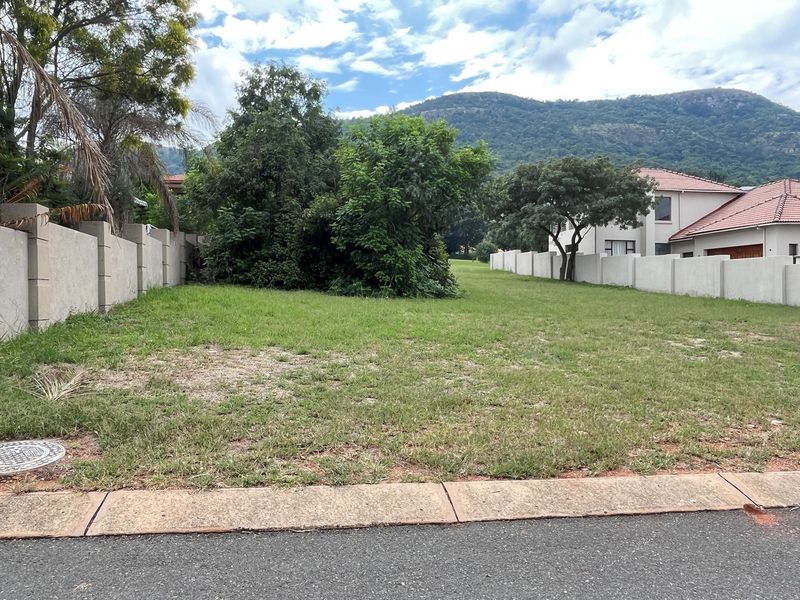 A vacant residential plot featuring a grassed surface, perimeter boundary walls, and a concrete curb, set against a scenic mountain backdrop.