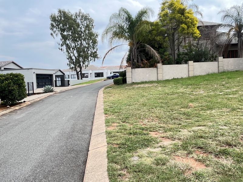 A view of the residential street approach featuring an asphalt driveway, concrete boundary wall, and established landscaping within the estate.