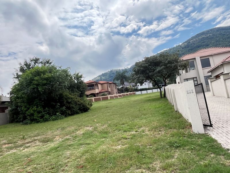 A vacant residential plot featuring a paved driveway, perimeter walling, and established landscaping with mountain views in the background.