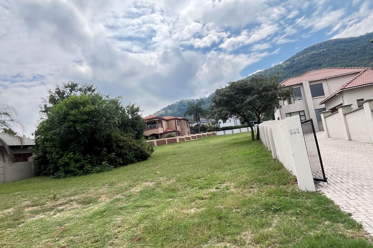A vacant residential plot featuring a paved driveway, perimeter walling, and established landscaping with mountain views in the background.