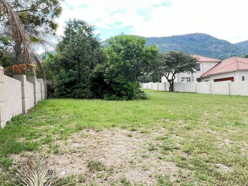 A spacious residential plot featuring a well-maintained grass lawn, perimeter boundary walls, and mature trees with scenic mountain views in the background.