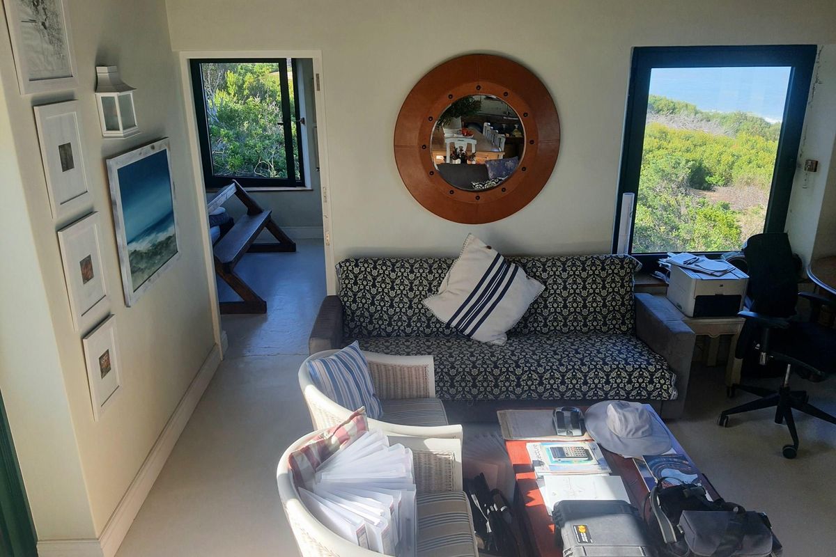 A living room area featuring large windows with natural light and views, connected to an adjacent room through a doorway.