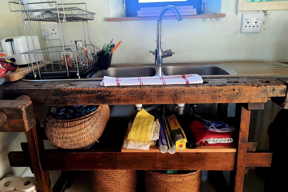 A rustic kitchen sink area featuring a double stainless steel basin set into a repurposed wooden workbench with open storage underneath.