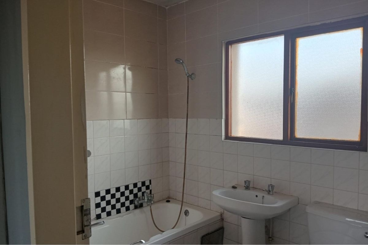 A dated bathroom featuring a standard bathtub with handheld shower attachment, pedestal basin, and toilet, finished with neutral wall tiling.