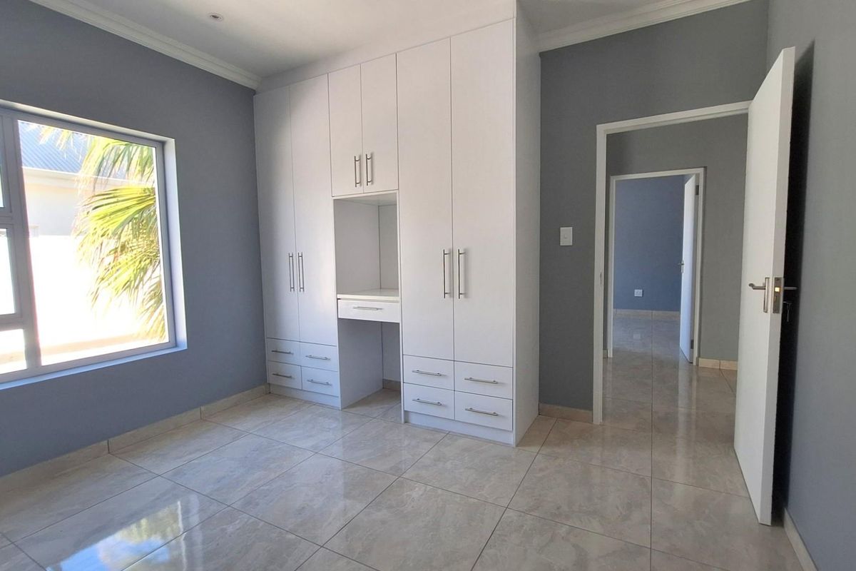 A bright bedroom featuring neutral grey walls, large porcelain floor tiles, and a modern white built-in wardrobe with an integrated vanity desk.