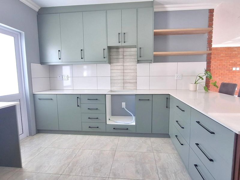 Modern kitchen featuring sage green cabinetry, white stone countertops, tiled backsplash, and an integrated breakfast bar with open shelving.